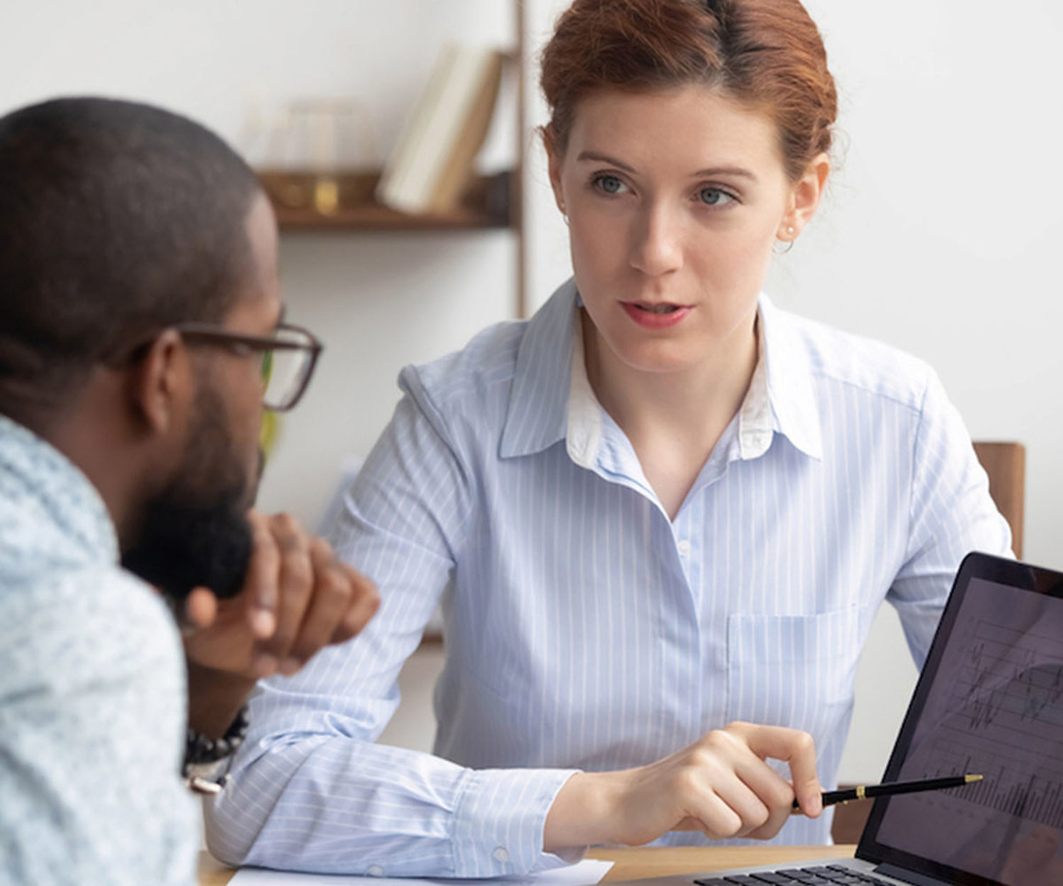Diverse colleagues analyzing diagrams together, using laptop, looking at screen, working on project, Caucasian businesswoman, mentor explaining business strategy to African American trainee (Diverse colleagues analyzing diagrams together, using laptop